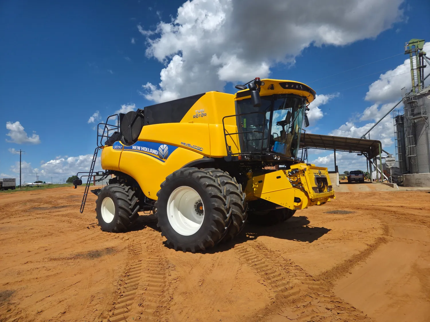 New Holland combine harvester beside grain silos
